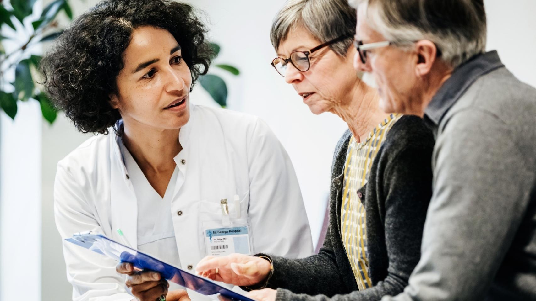 Woman doctor sitting down and showing a medical chart to an elderly couple.
