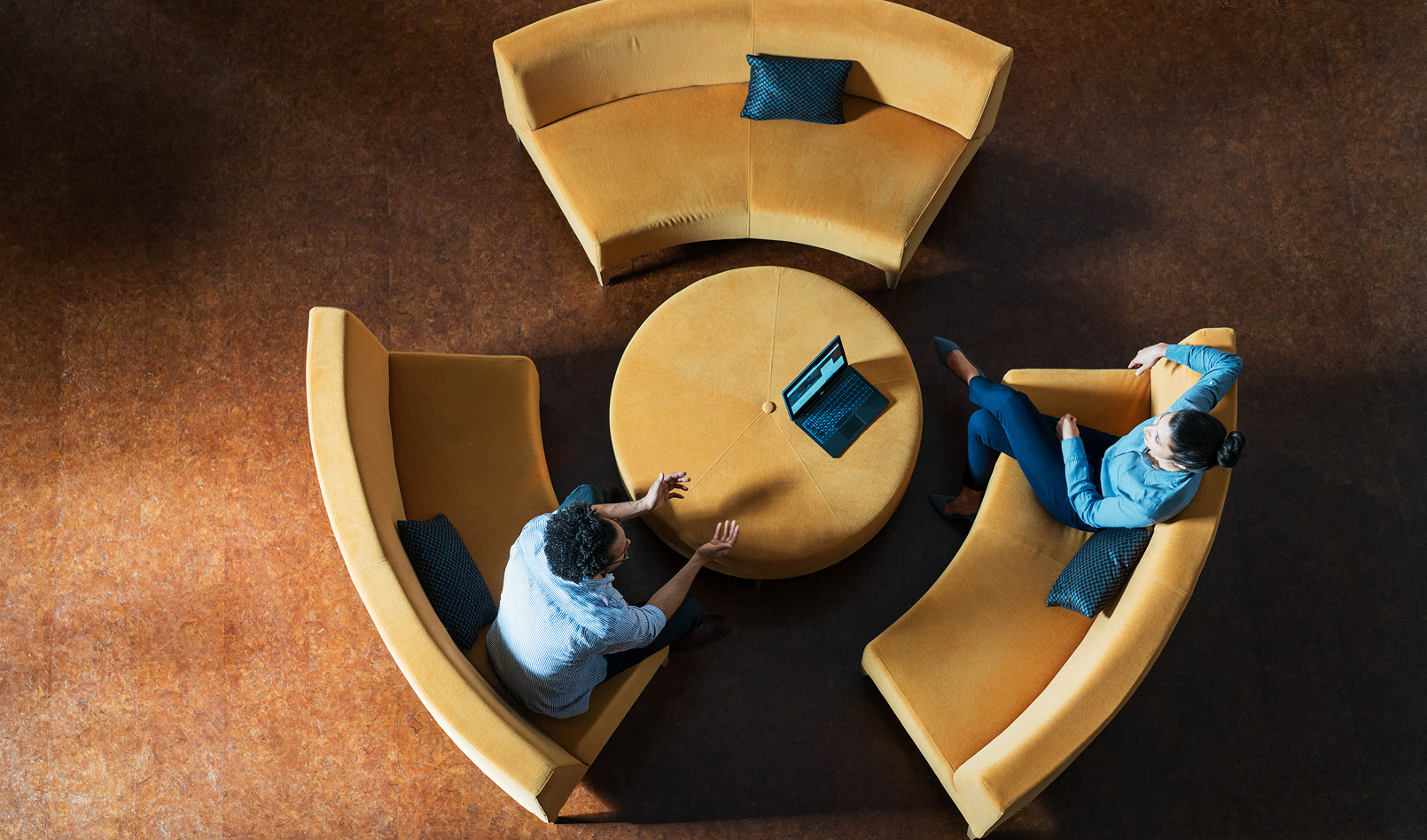 An overhead view of two professionals sitting in a modern lounge area around a circular table with curved benches. They're facing each other, engaged in conversation, with a laptop on a central table.