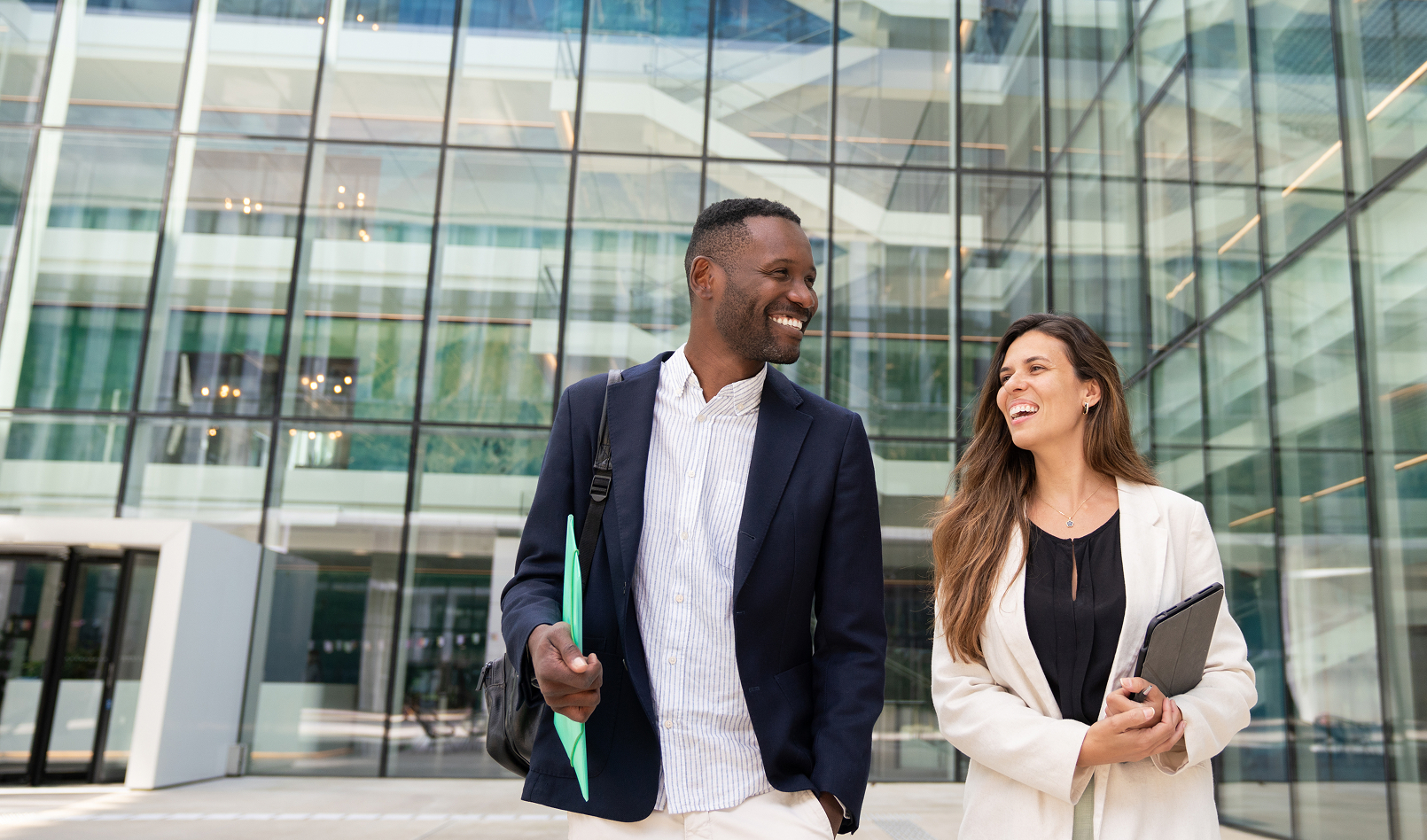 Two professionals, a man and a woman, are smiling and walking confidently in front of a modern glass office building. They represent the engaging opportunities and collaborative environment available in careers at Blue Harven .