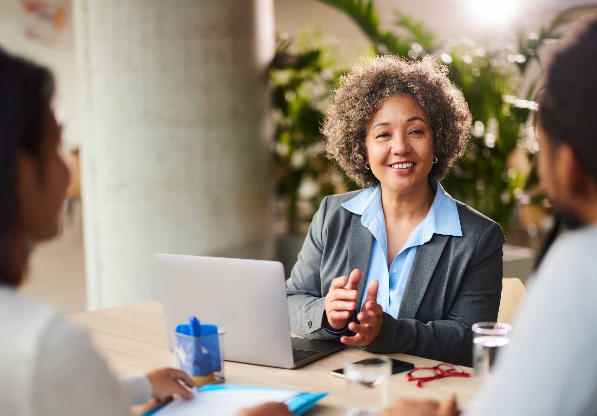 A Blue Harven Bank financial advisor with short curly hair is smiling and gesturing while seated at a table with her computer. She is meeting with a client to discuss their financial needs, symbolizing the expert and personalized service we offer.
